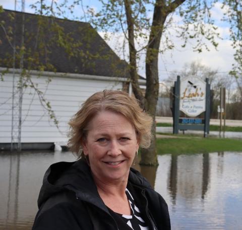 Leah Rasmussen stands on a dry portion of Highway W with floodwaters and her shop and home in the background. 