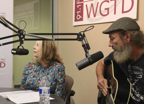 Jill Plaisted sits in front of a mic at WGTD and Keith Stokes is strumming a guitar. 