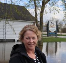 Leah Rasmussen stands on a dry portion of Highway W with floodwaters and her shop and home in the background. 